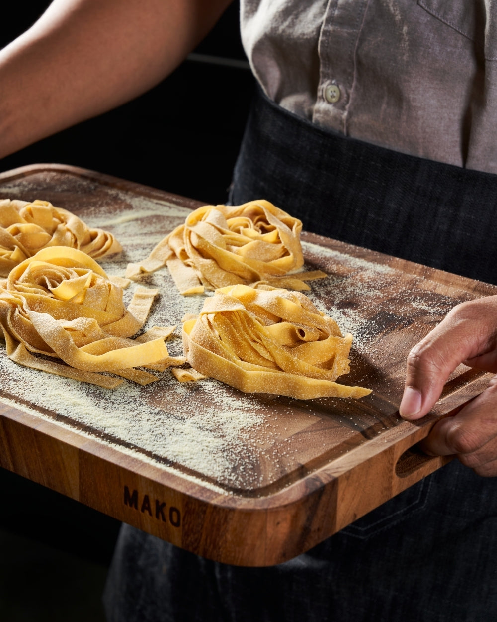 Wood chopping board with fresh pasta nests and flour, held by person in apron
