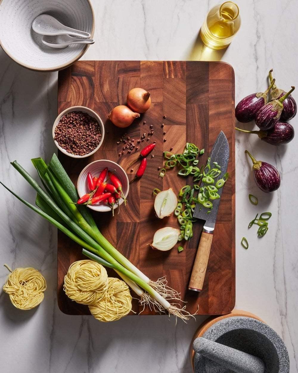 Large wooden chopping board with chef knife, spring onions, chili, noodles, onion, and spices.