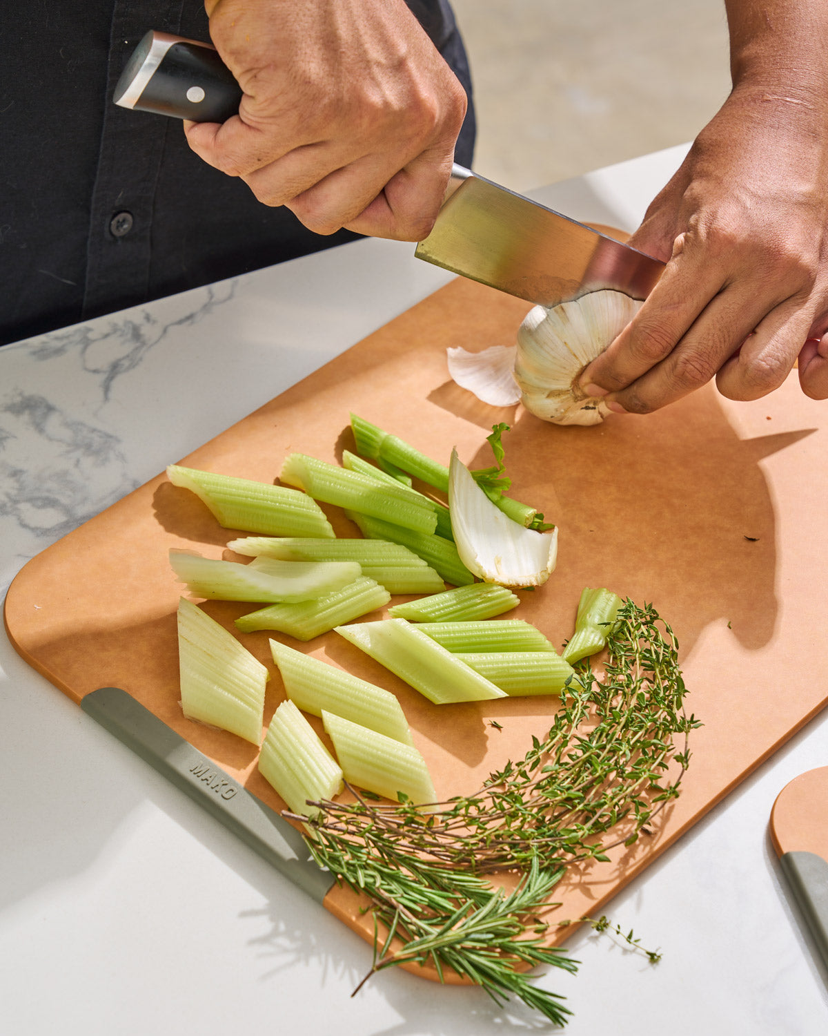 Wood fibre non-slip chopping board with celery, fresh herbs, and garlic being sliced