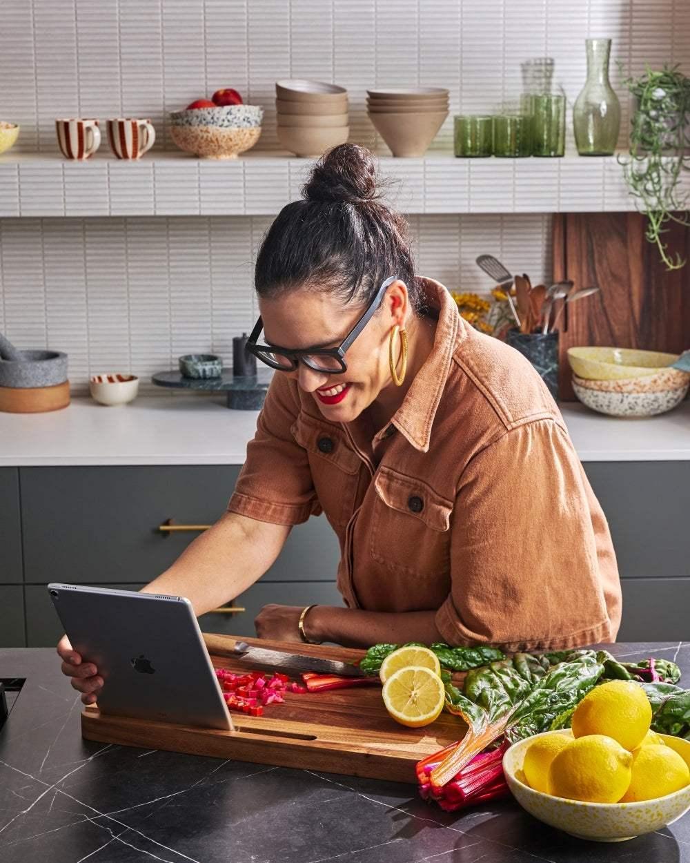 Woman in brown jacket using tablet and chopping vegetables on wooden cutting board in modern kitchen