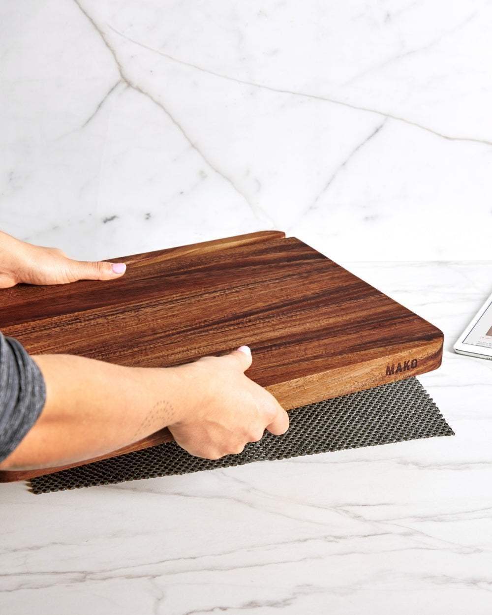 Hands placing a Mako wooden chopping board on a kitchen counter with marble backsplash