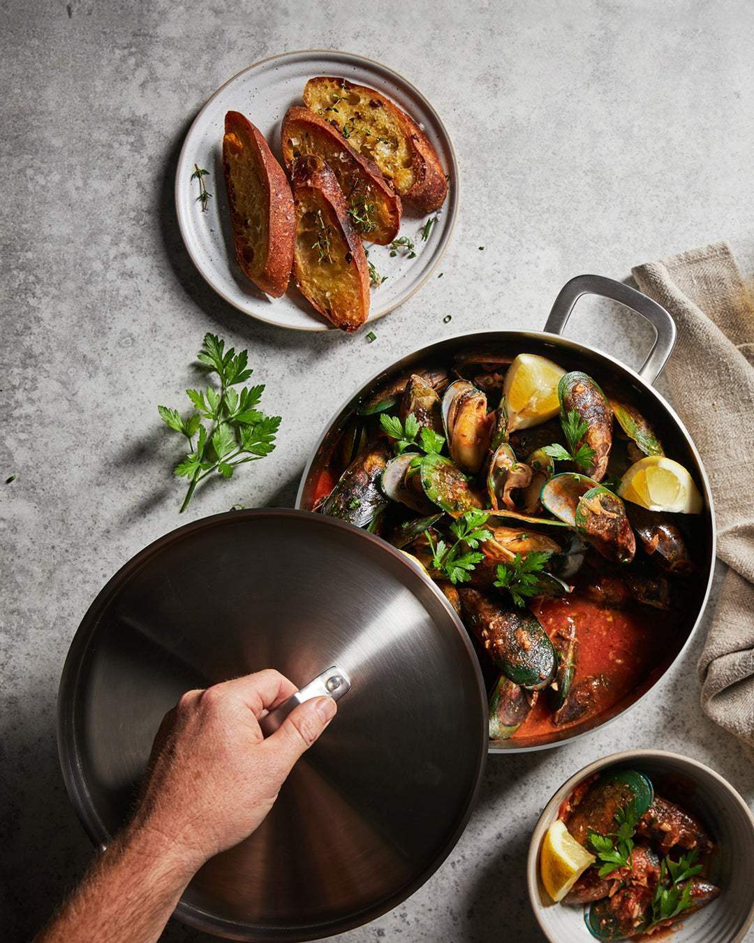 Mako 28cm Genius 5-ply essential pan with lid holding cooked mussels in tomato sauce, lemon wedges, and parsley, next to toasted garlic bread on plate