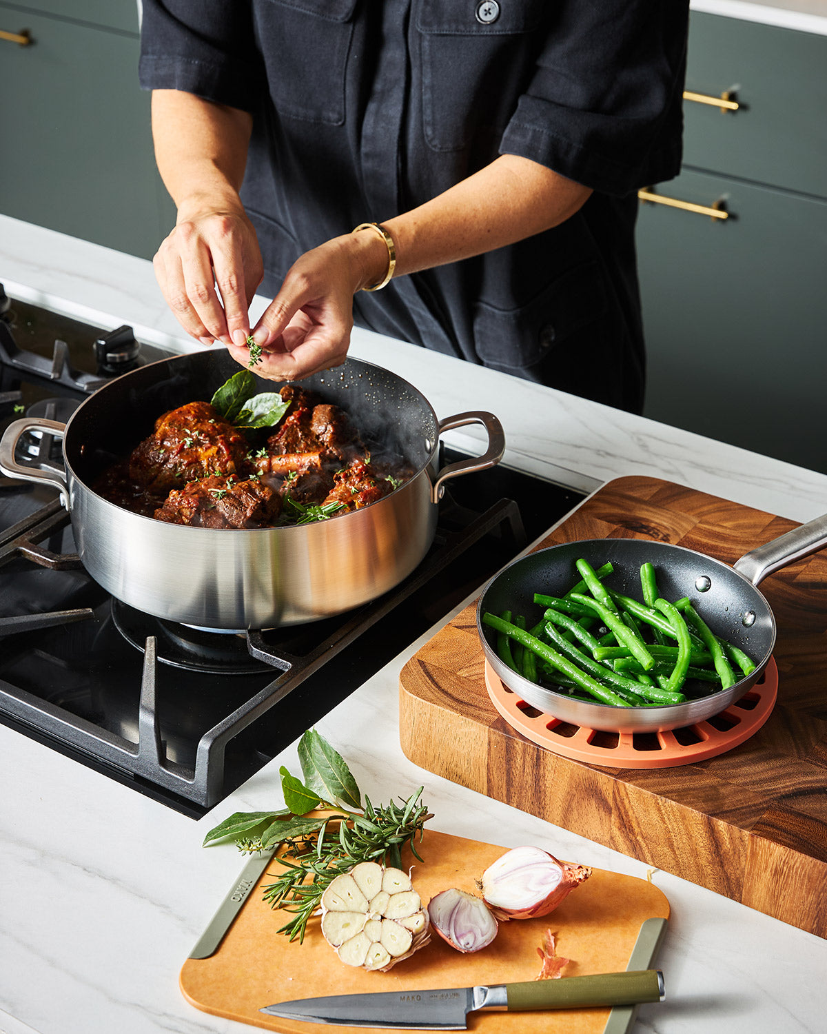 Person garnishing meat stew in stainless steel non-stick pan on stovetop, with green beans in skillet nearby, fresh herbs and garlic on cutting board