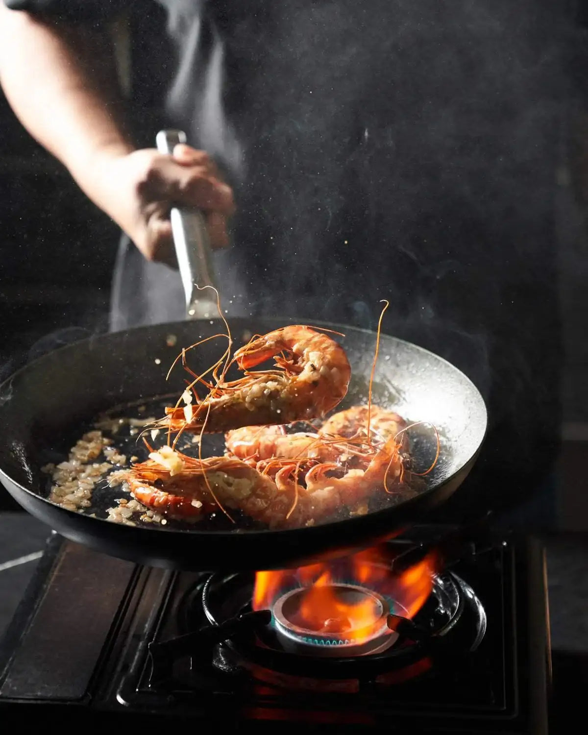 Mako black steel frying pan with prawns sautéing on gas stove flame, cooking with garlic