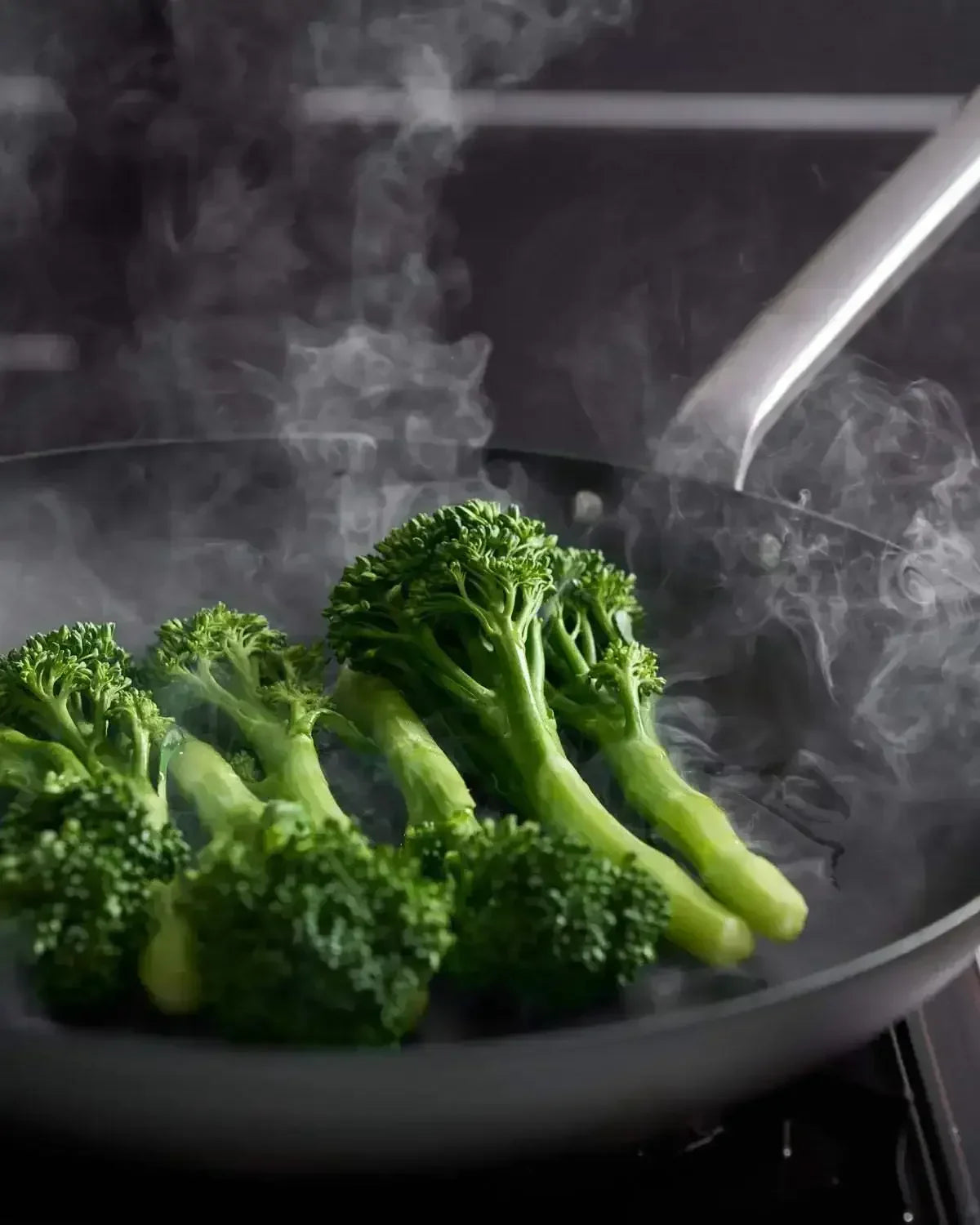 Fresh broccolini steaming in a black steel frying pan on a stovetop