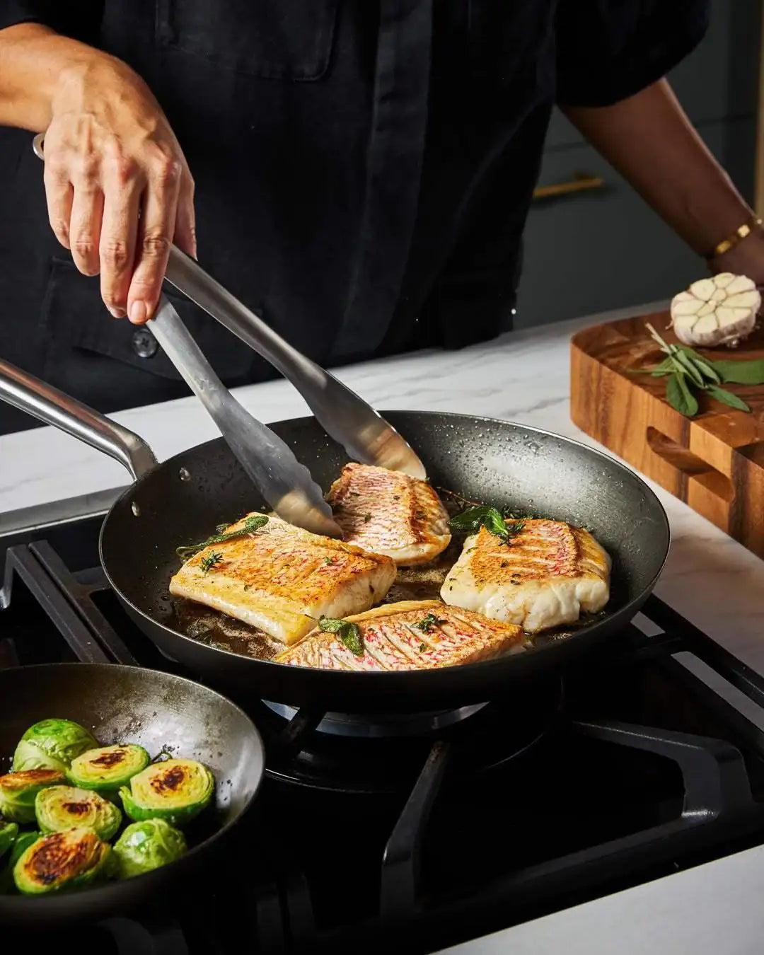 Hand using tongs to cook golden fish fillets with herbs in a Mako black steel frying pan on a gas stove