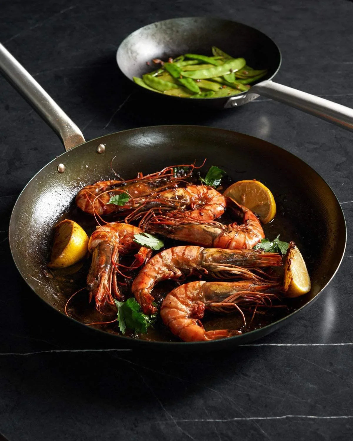 Black steel frying pans with cooked shrimp and lemon wedges, and stir-fried green beans on dark surface