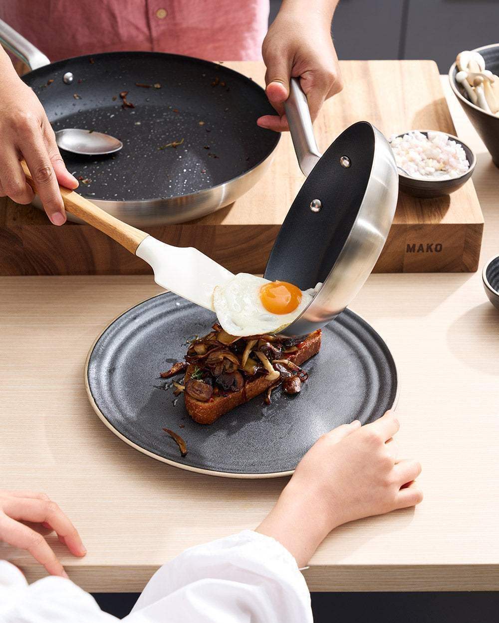 Hands using Mako non-stick frying pan to serve fried egg on mushroom toast, kitchen setting with cutting board