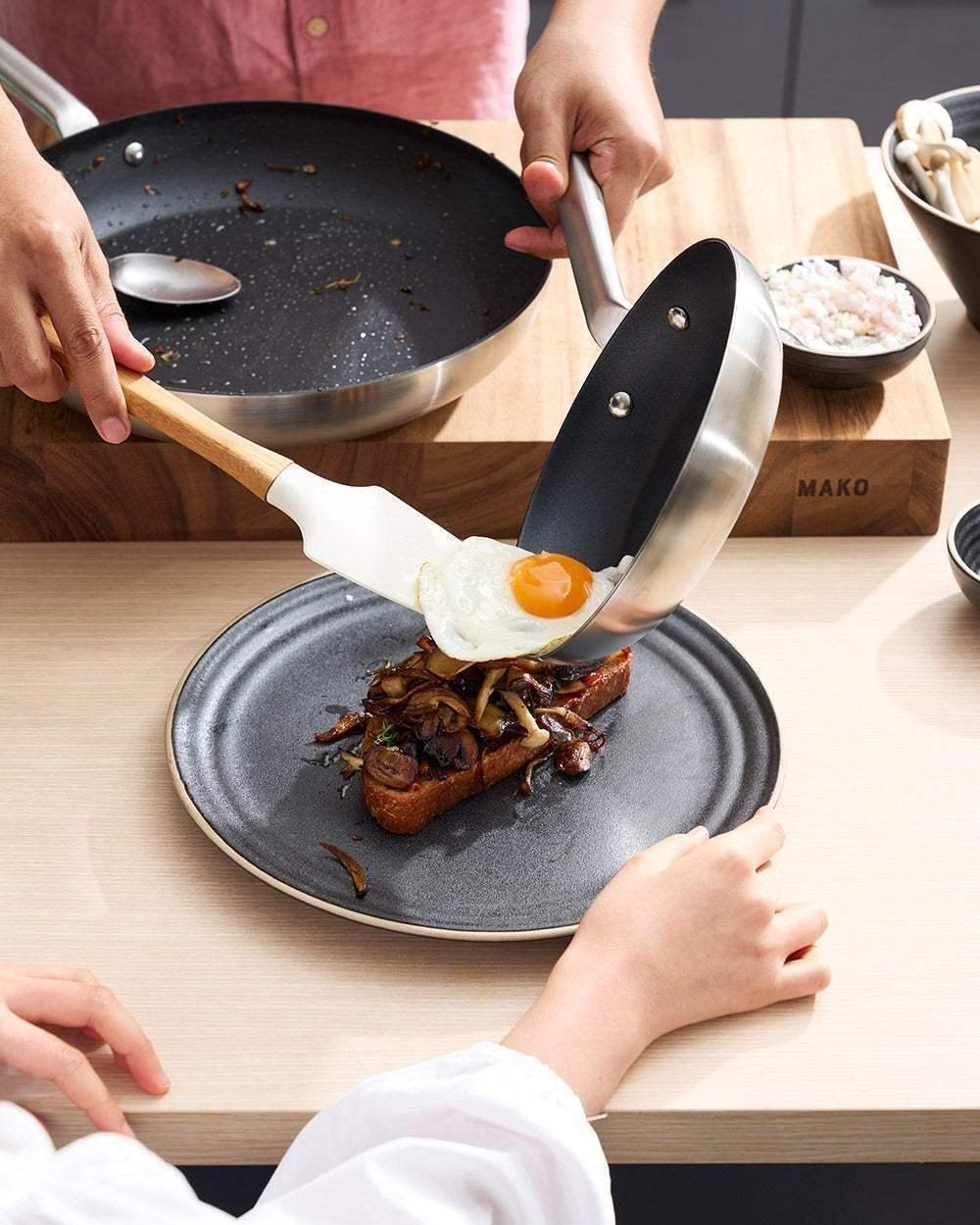 Non-stick frying pan serving fried egg onto toast with mushrooms on a ceramic plate