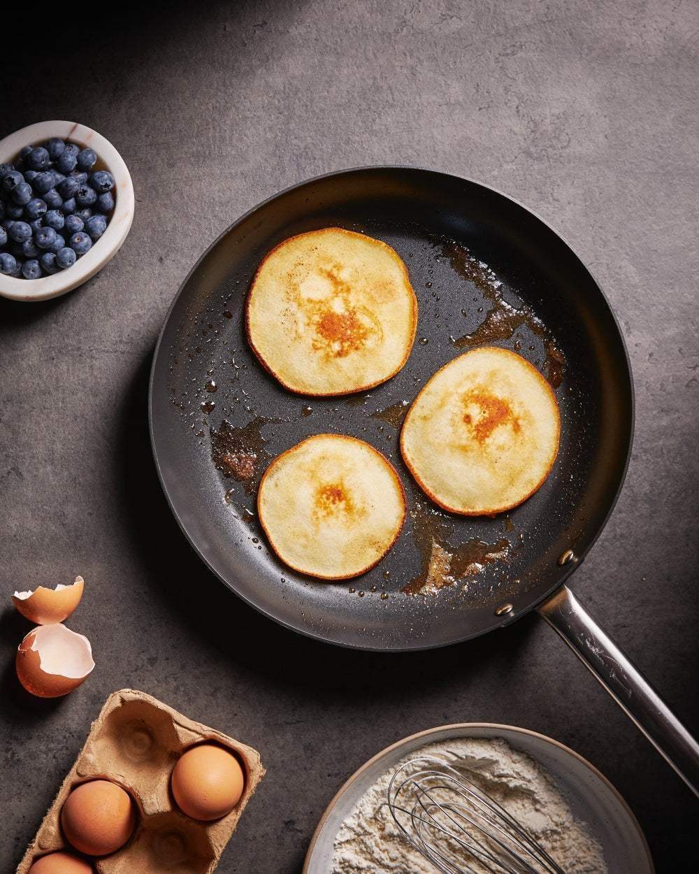 Non-stick Mako frying pan with three golden pancakes cooking, surrounded by eggs, whisk, flour, and bowl of blueberries
