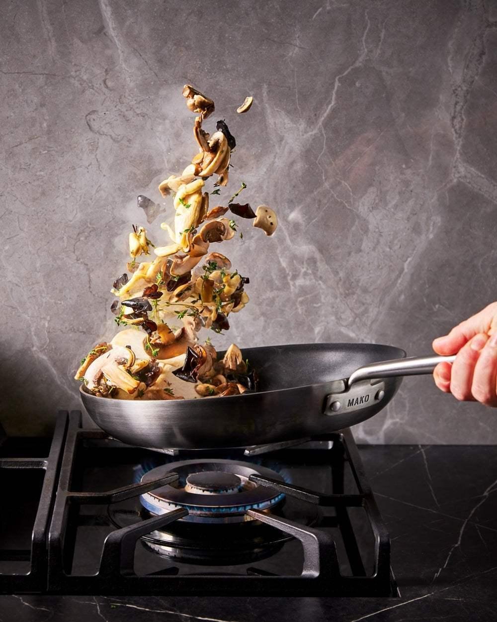 Hand flipping mushrooms and herbs in a Mako non-stick frying pan on a gas stove with marble backsplash