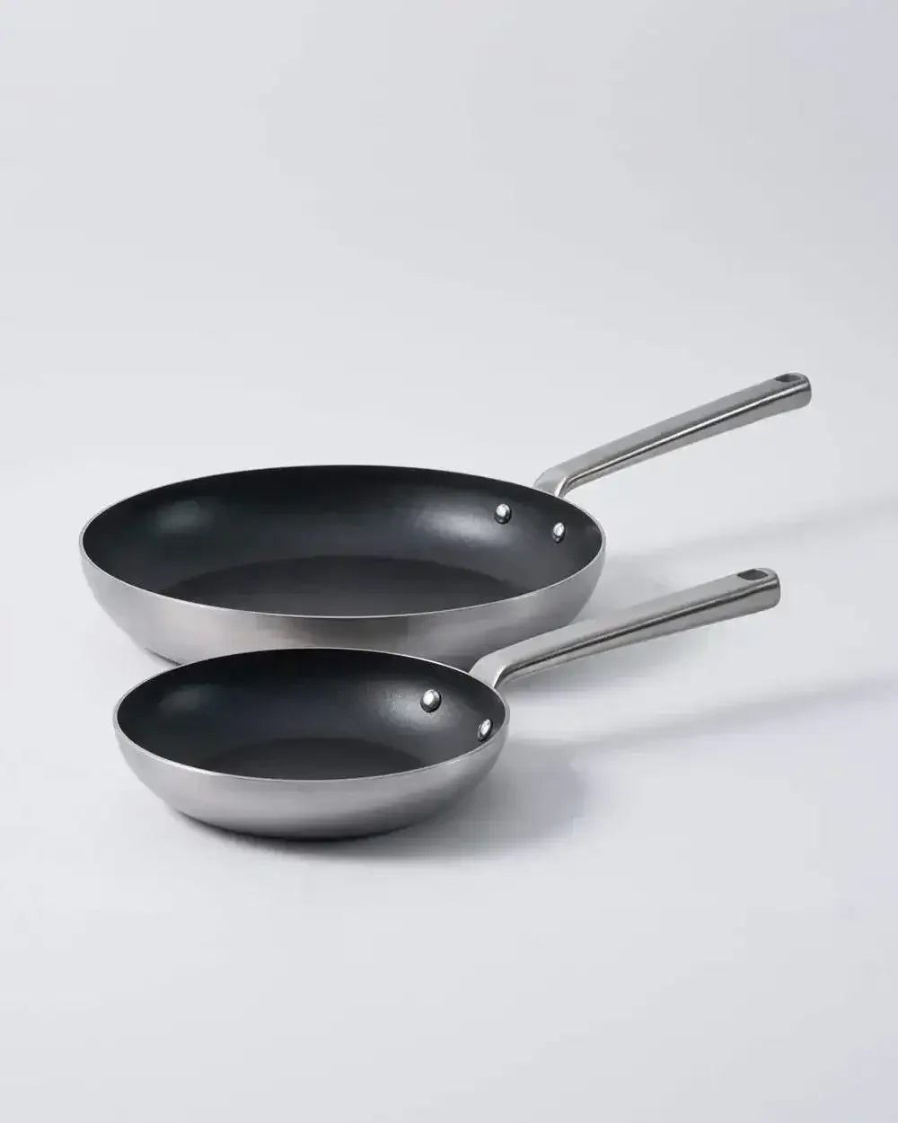 Two stainless steel non-stick frying pans with metal handles on a light background