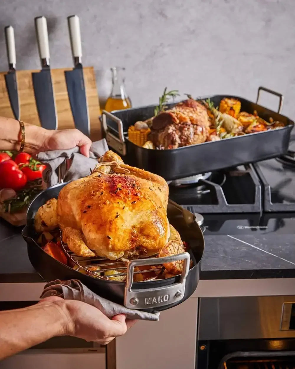 Roast chicken in MAKO non-stick roasting pan with kitchen knives and cooked roast in background