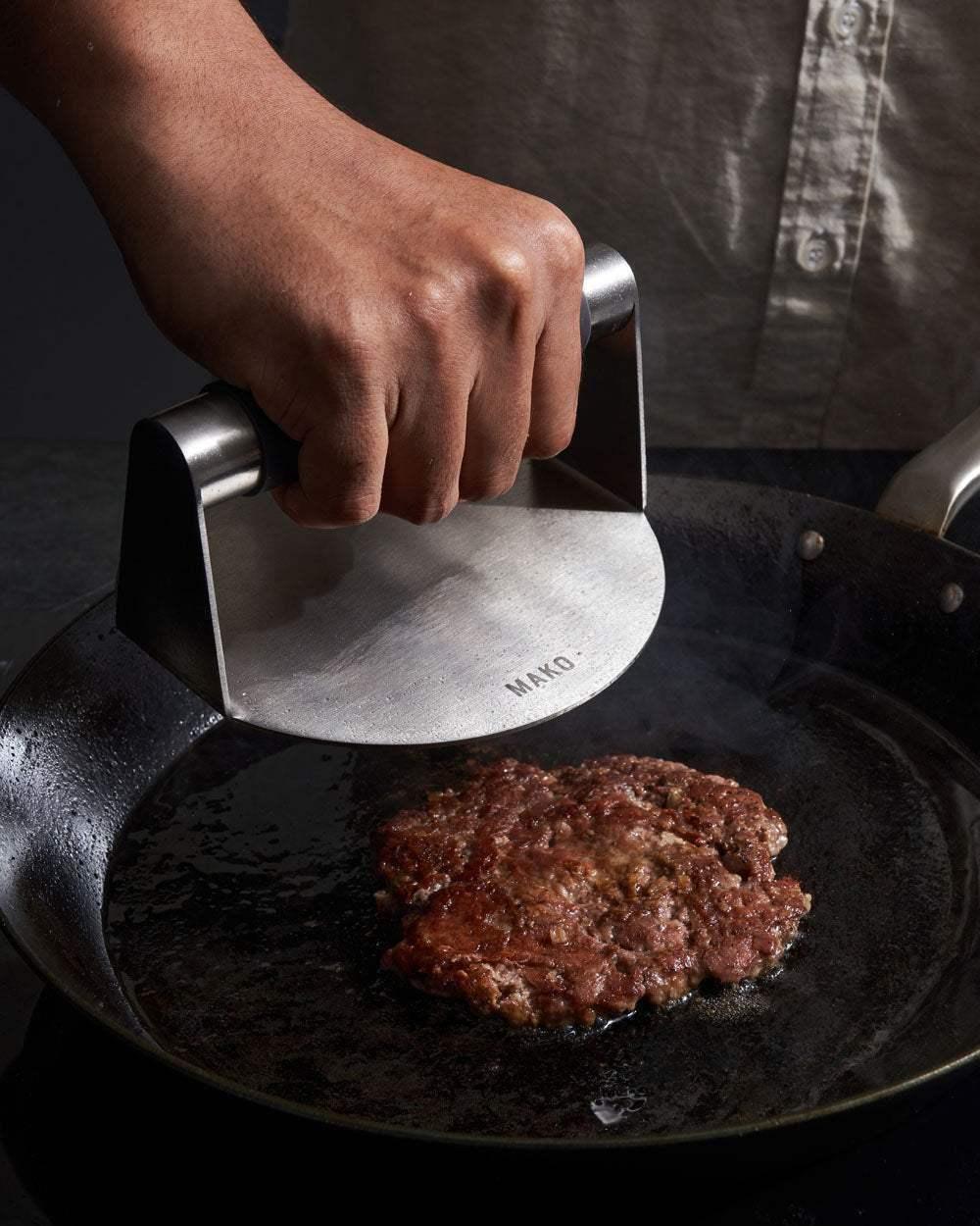 Hand pressing a burger patty on a skillet with a stainless steel burger press