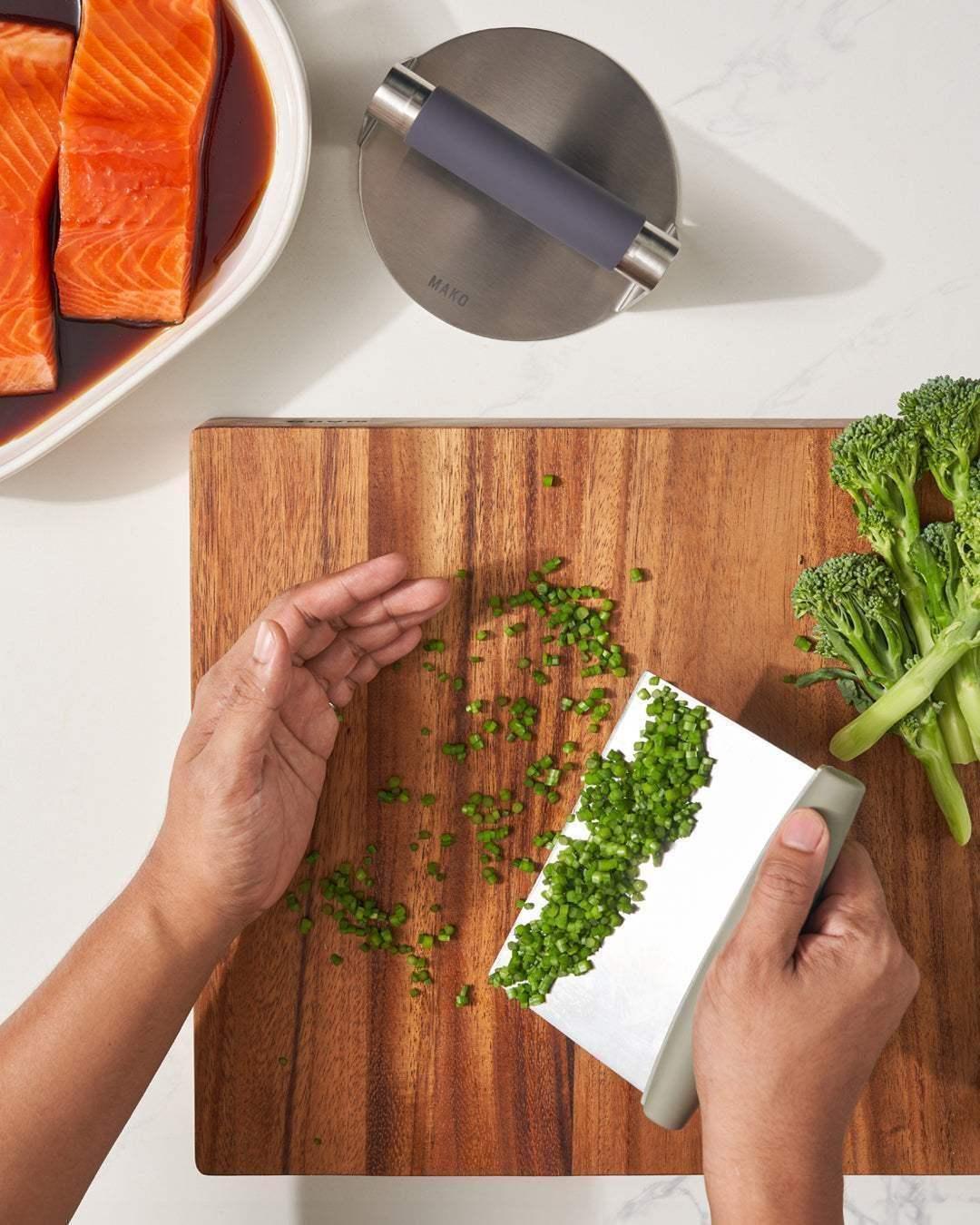 Hand using a kitchen scraper to gather chopped green onions on a wooden cutting board, with broccoli and marinated salmon nearby.