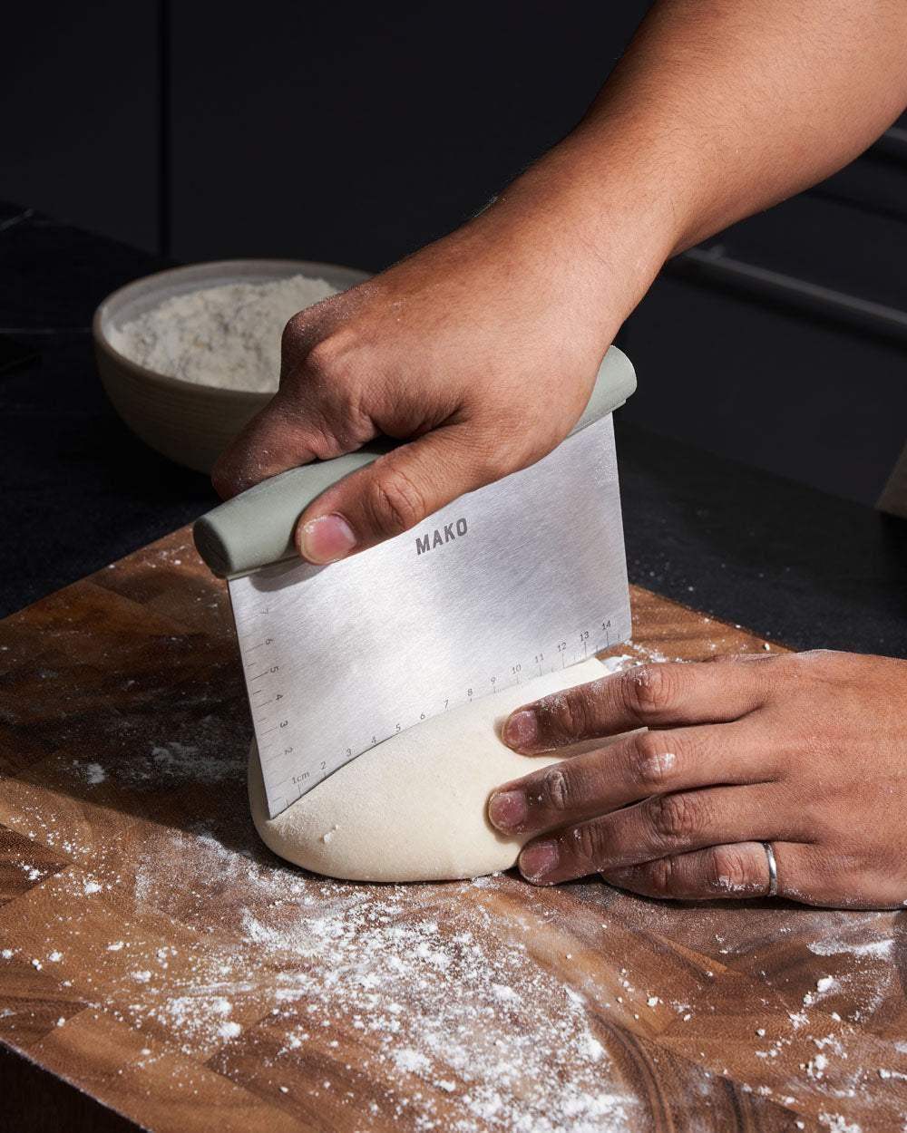 Hands using Mako dough scraper to cut flour-dusted dough on wooden board with bowl of flour in background