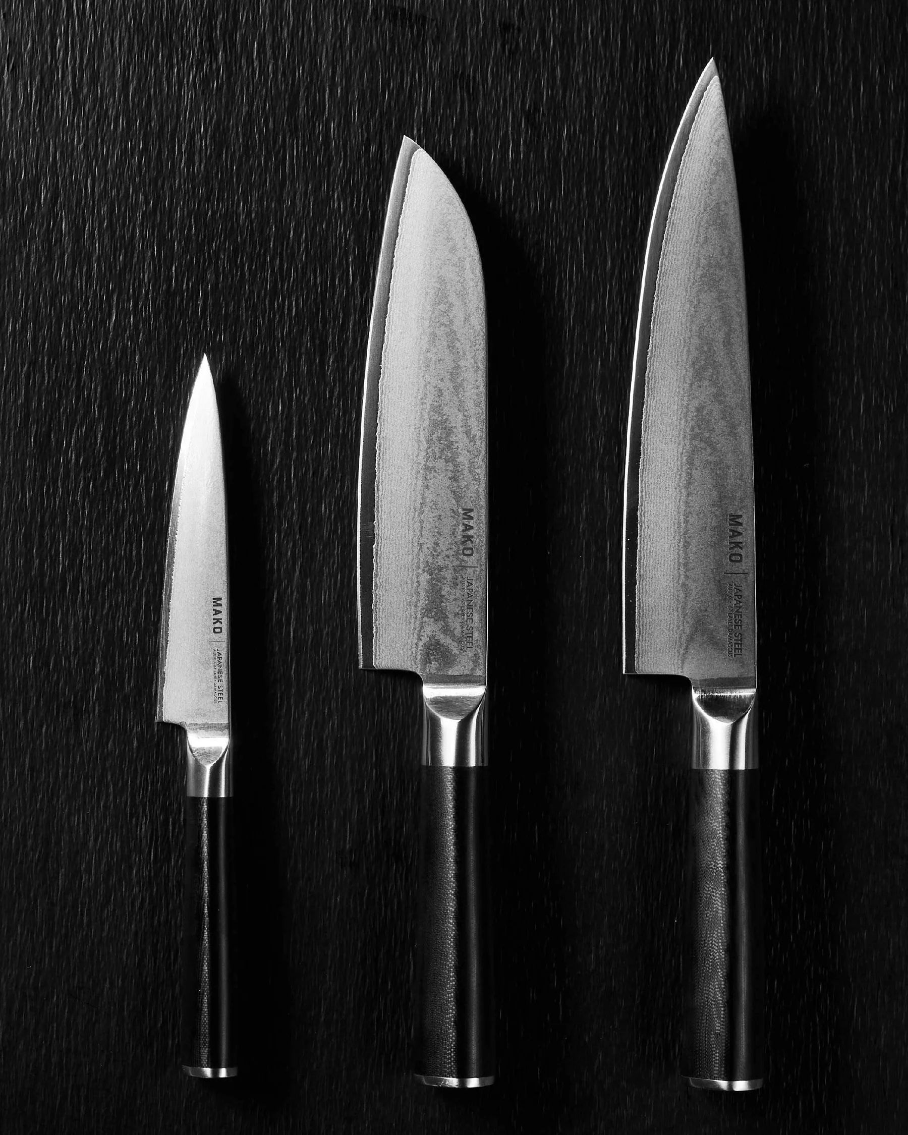 Three Japanese steel kitchen knives with black handles on a dark textured surface.