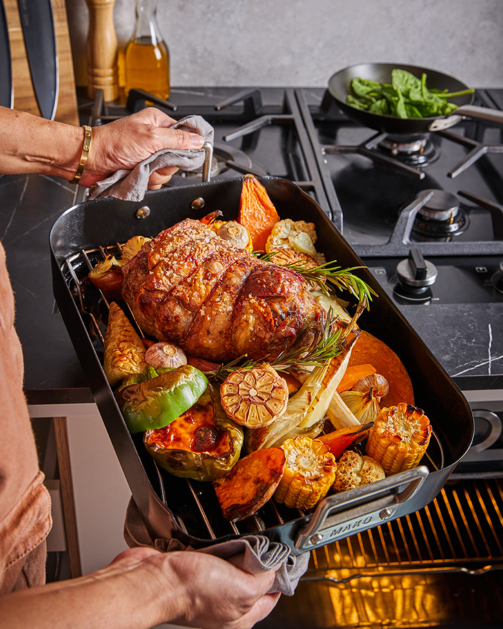 Person holding non-stick roasting pan with roast meat, vegetables, and herbs by stovetop