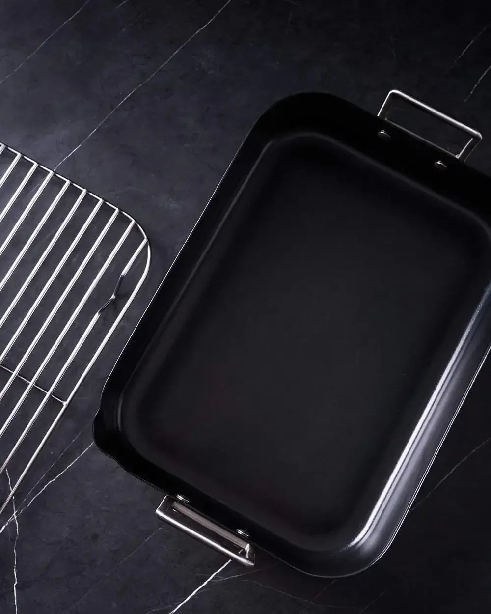 Non-stick roasting pan with metal handles next to a stainless steel rack on a dark countertop