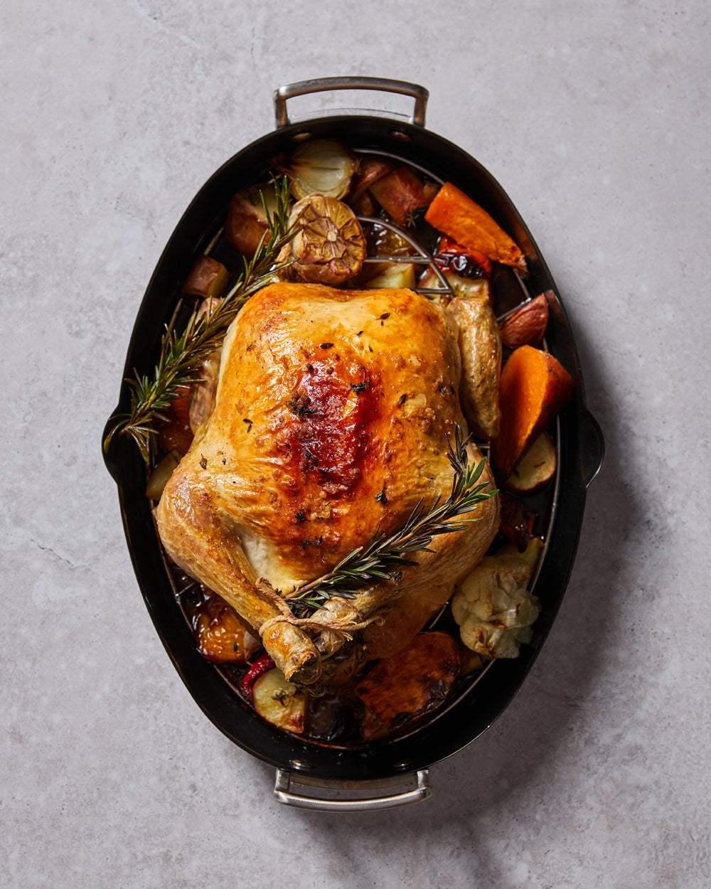 Oven-roasted whole chicken with crispy skin, herbs, and assorted roasted vegetables in a black oval roaster