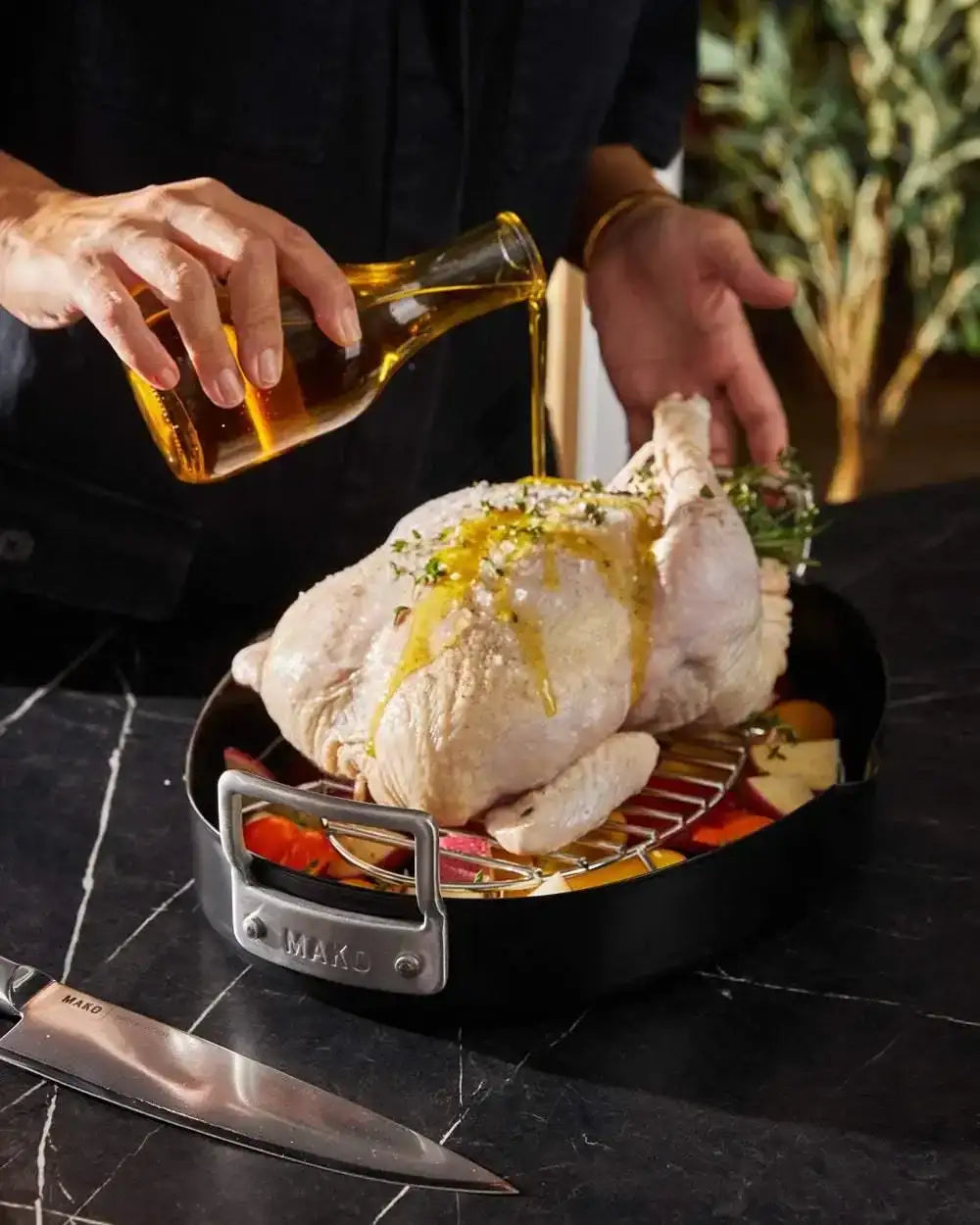 Raw whole chicken in Mako non-stick oval roaster being drizzled with oil, with vegetables underneath and chef's hands visible
