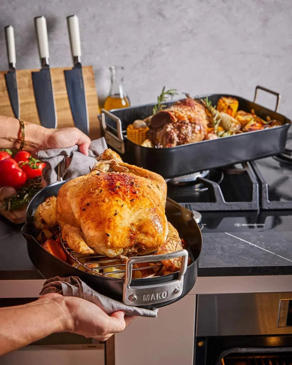 Hands holding a Mako non-stick roaster with roasted chicken on a kitchen countertop with knives and cooked meat in the background