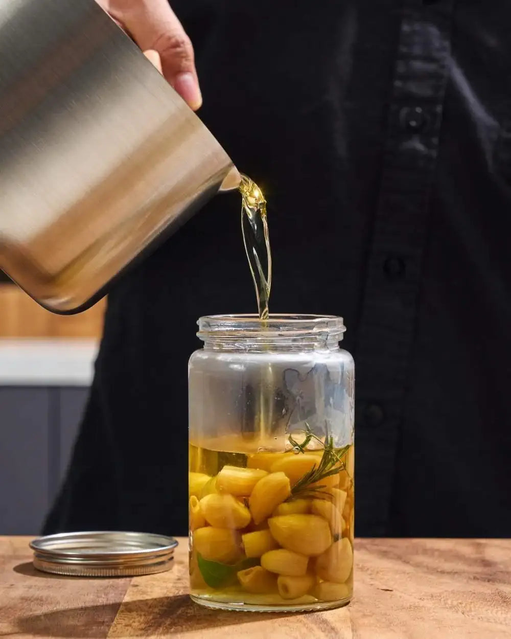 Pouring oil from a brushed steel Mako saucepan into a jar with garlic and herbs, kitchen setting