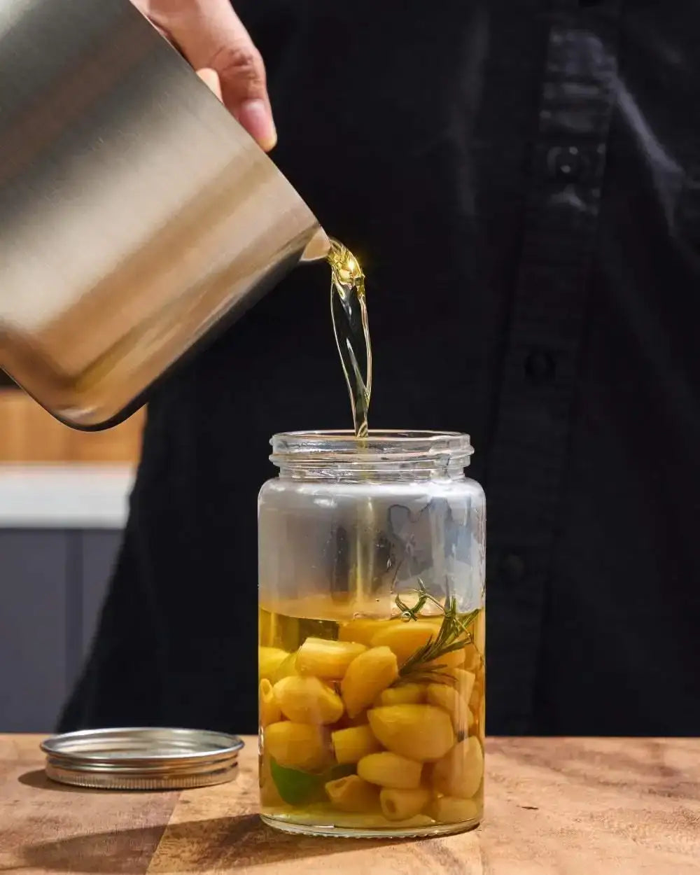 Stainless steel saucepan pouring oil over garlic and herbs into a glass jar on a kitchen counter