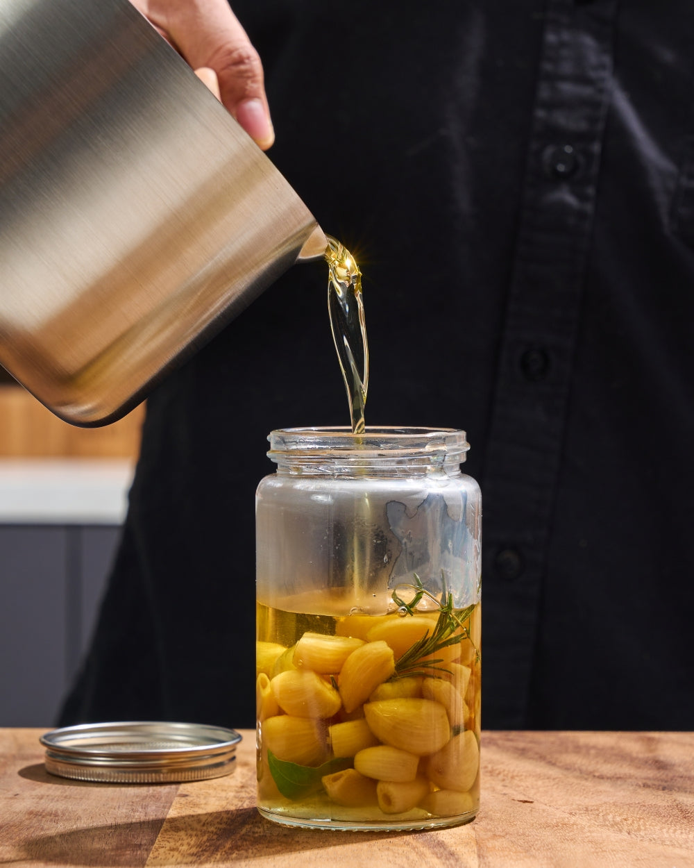 Stainless steel saucepan pouring oil over garlic and herbs into a glass jar on a kitchen counter