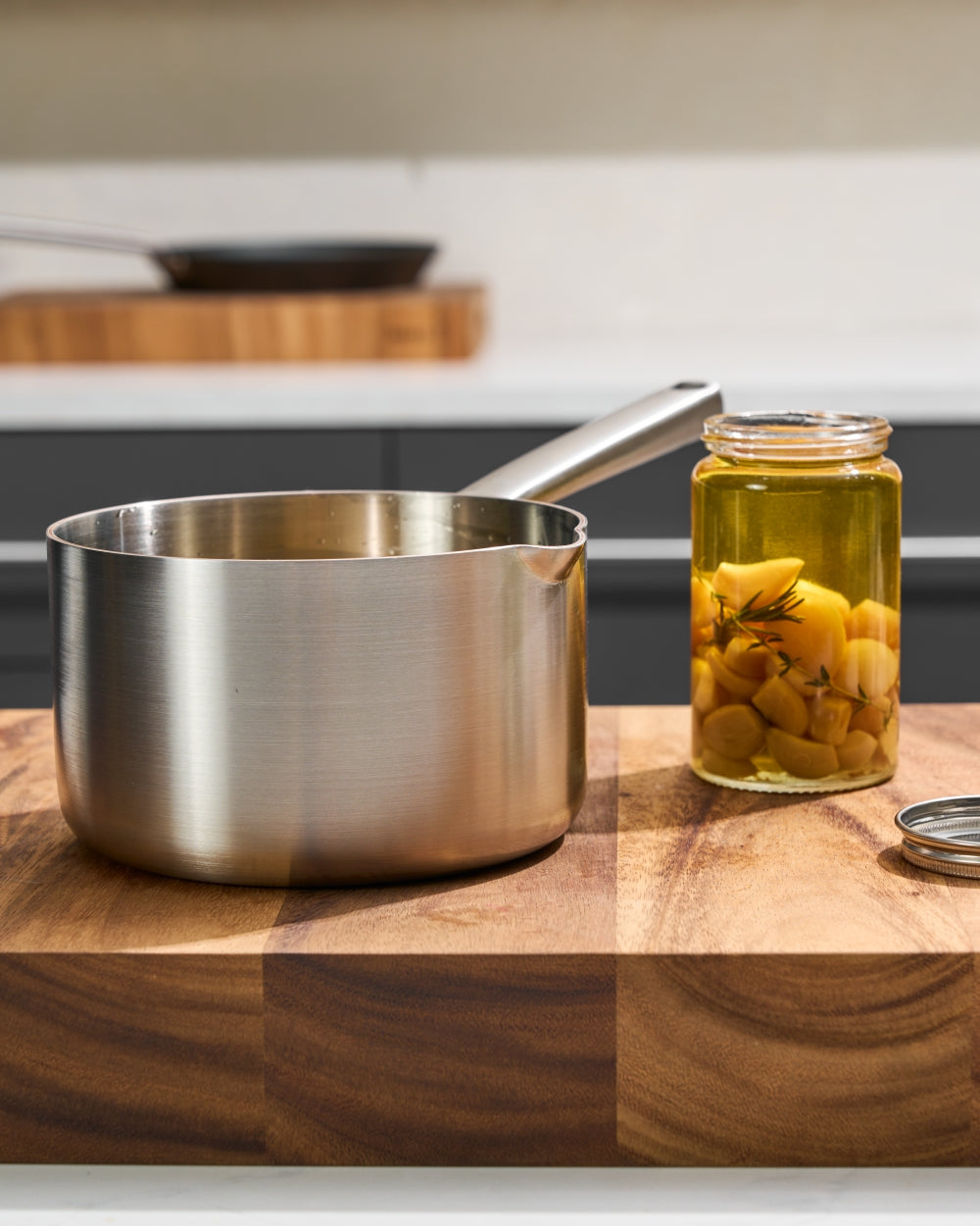 Stainless steel saucepan next to glass jar of preserved vegetables on wooden cutting board
