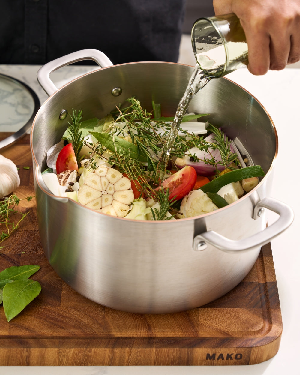 Stainless steel saucepan with fresh vegetables, herbs, and liquid being poured in