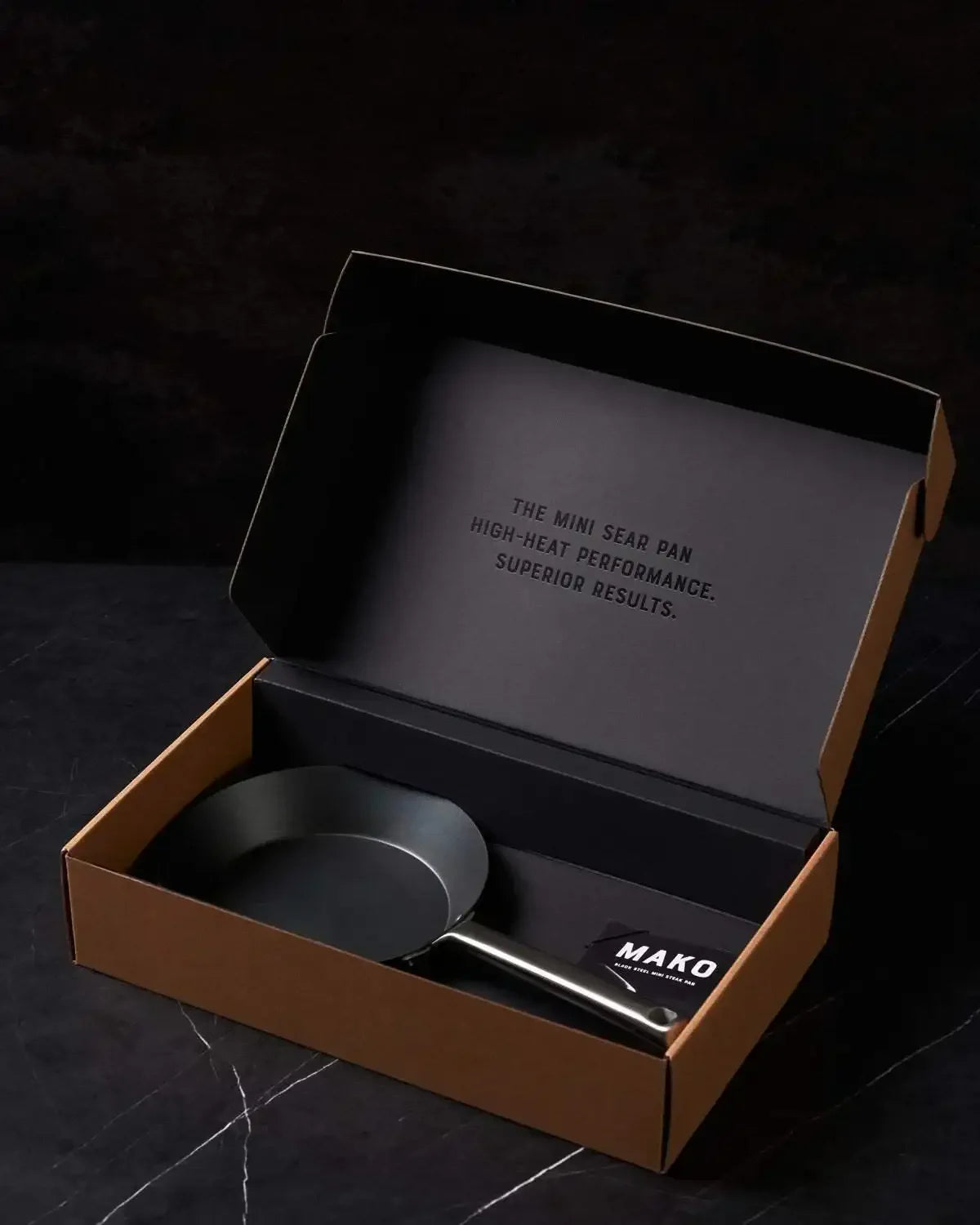 Mini black steel sear pan in open box on dark marble surface, premium cookware gift packaging
