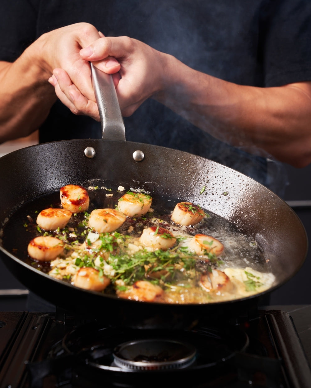 Searing scallops with herbs in a black steel steak pan on a stovetop