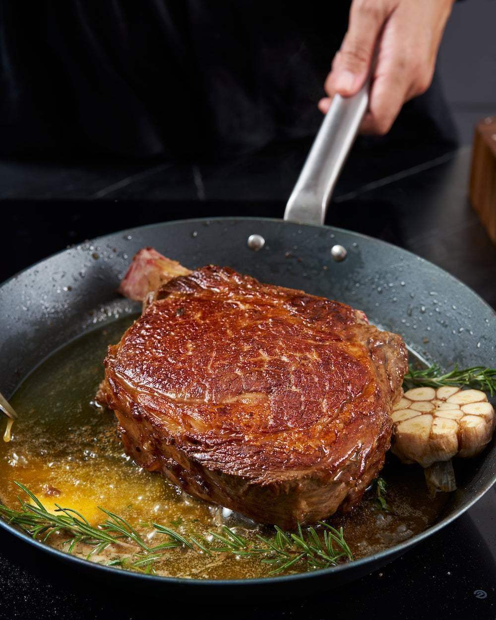 Thick steak searing in Mako black steel pan with garlic and rosemary, hand holding pan handle