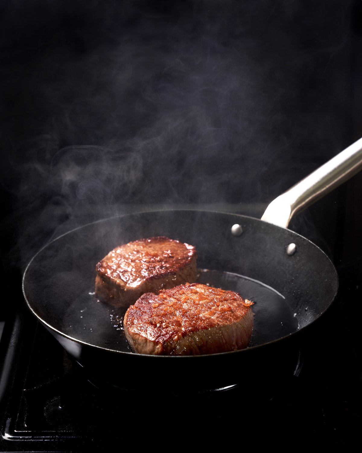 Two seared steaks cooking in a black steel frying pan on a stovetop