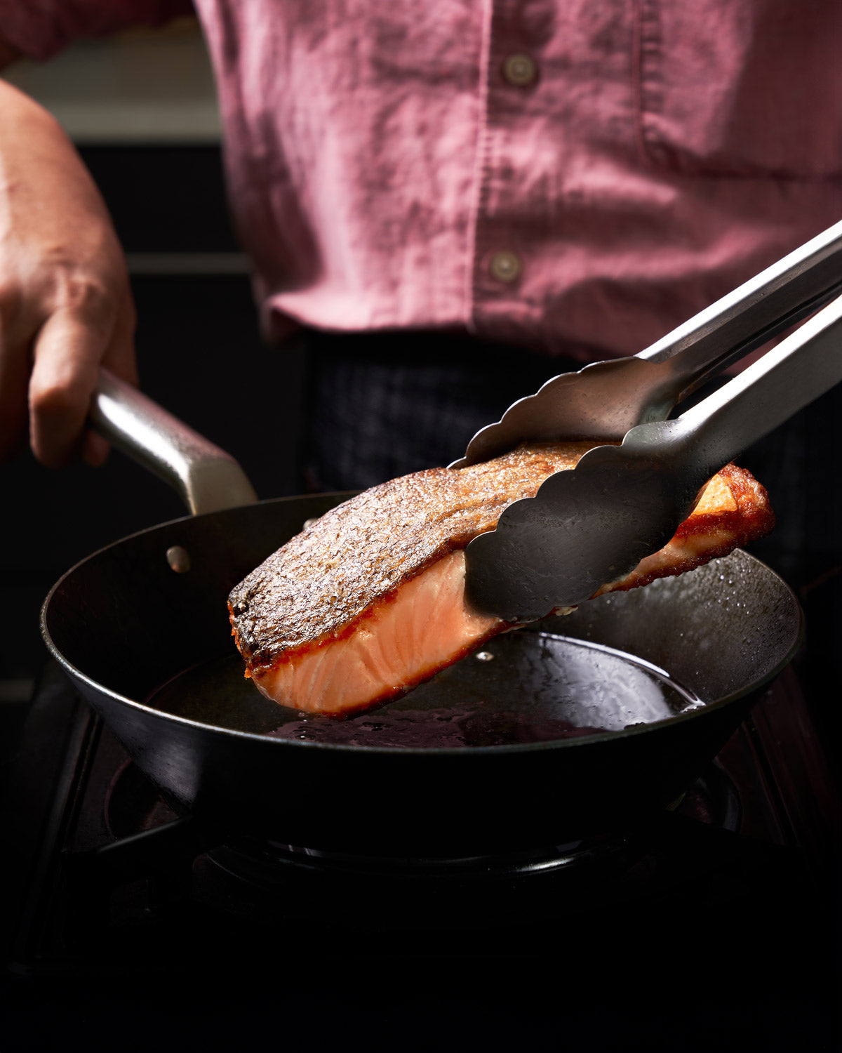 Person searing salmon steak in black steel pan with tongs on stovetop