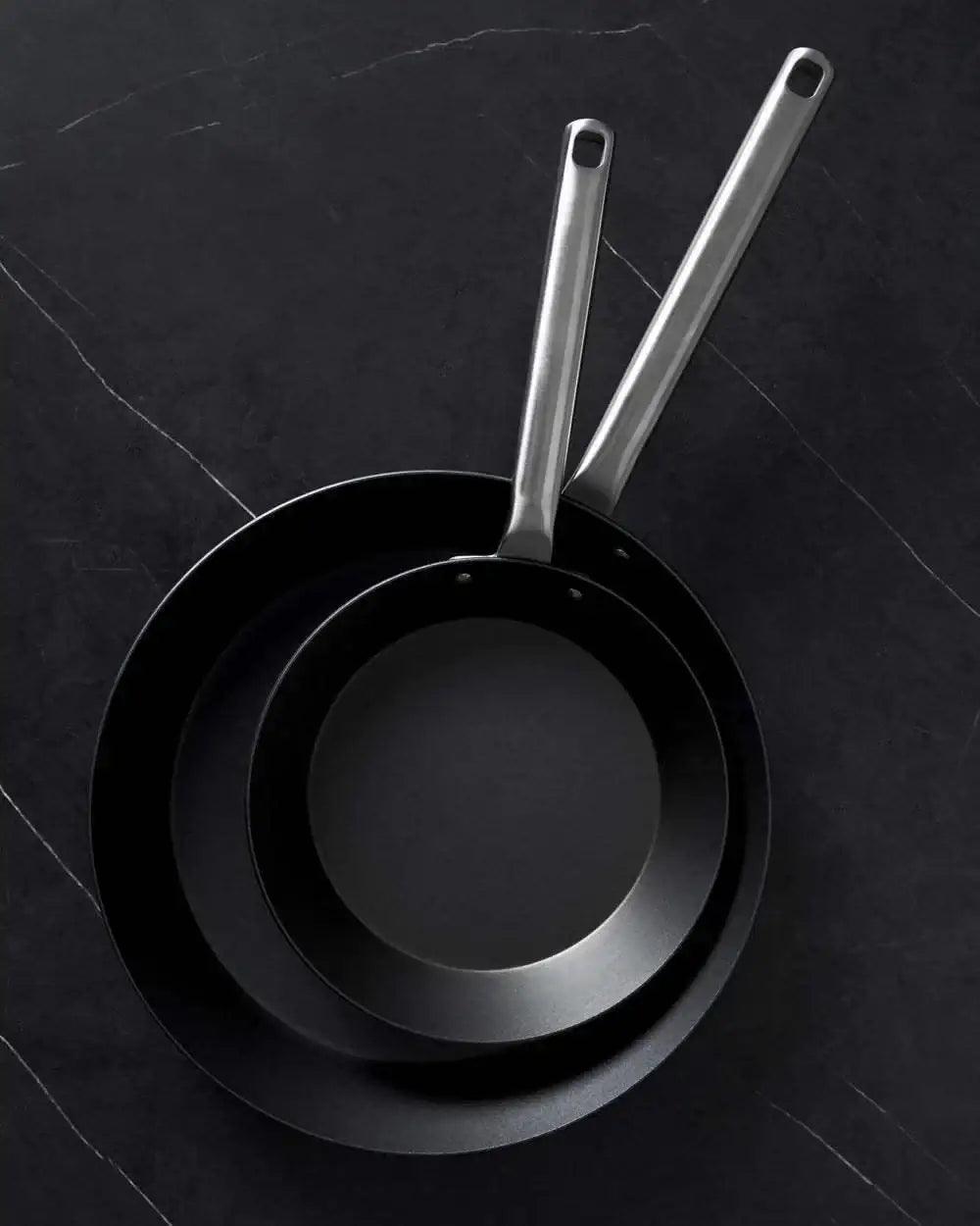 Two black steel frying pans with stainless steel handles on dark textured surface, Mako by Marion Grasby