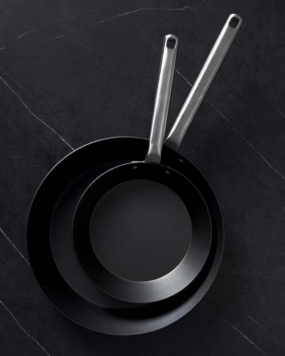 Two black steel frying pans with stainless steel handles on dark textured surface, Mako by Marion Grasby