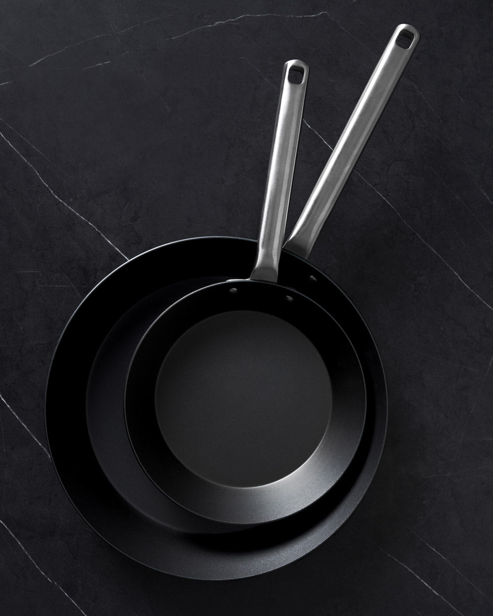 Set of two black steel steak pans with silver handles on a dark stone surface