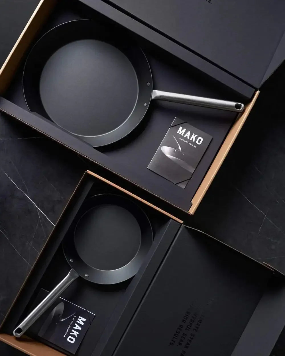 Black steel steak pan set with silver handles in gift boxes on dark surface