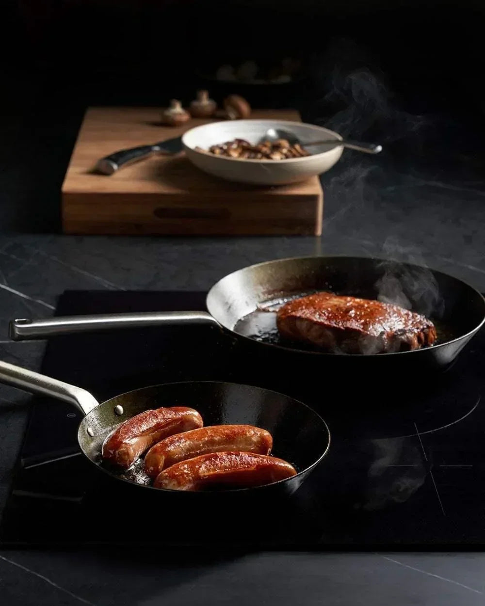 Two black steel pans cooking sausage and steak on a dark stovetop with mushrooms on a wooden cutting board