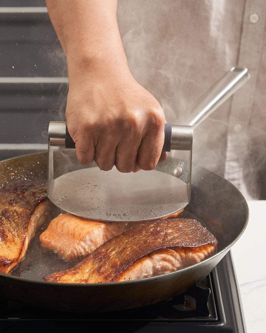Person using a metal steak press on searing salmon fillets in a black steel pan