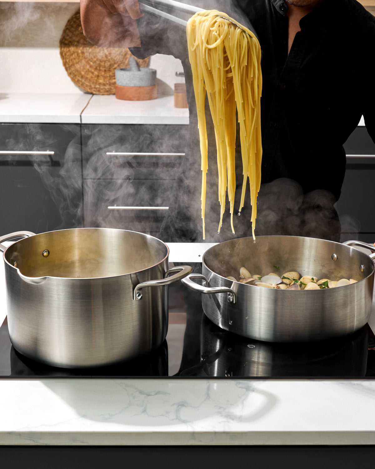 Stainless steel stock pot on stovetop next to sauté pan, pasta being lifted with tongs