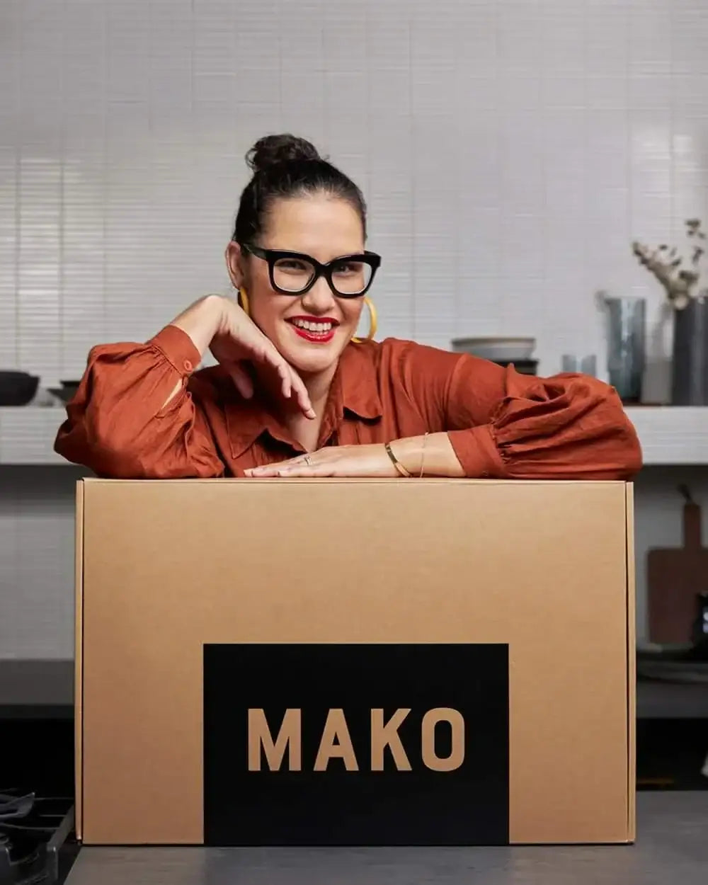 Woman with glasses in orange shirt leaning on large brown box with MAKO logo in modern kitchen