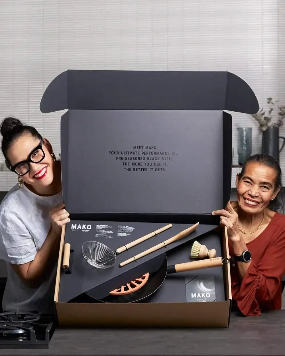 Two smiling women showing open Mako black steel wok essentials set box with utensils, endorsed by chef Marion Grasby