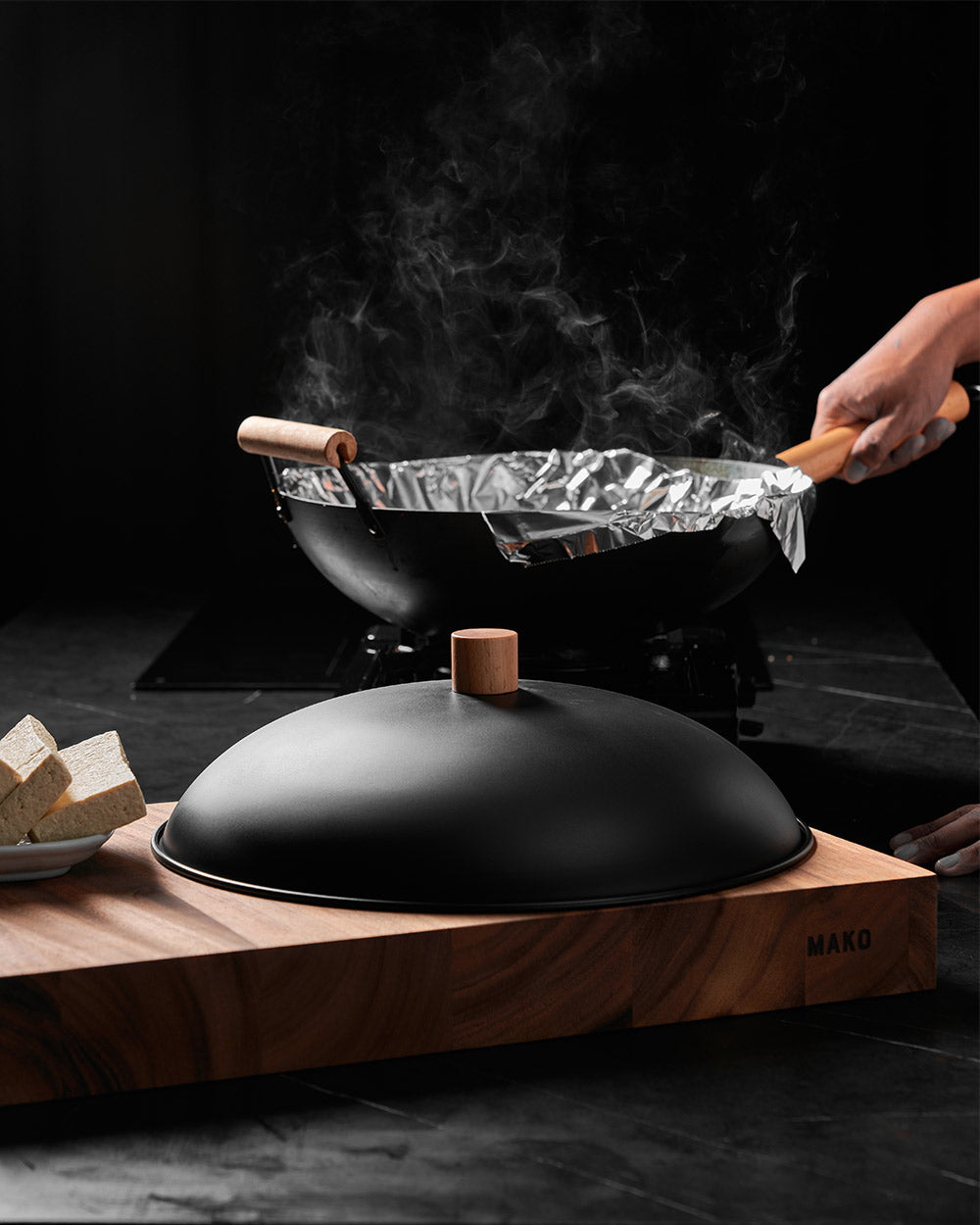 Black steel wok with wooden handle, matching lid, tofu cubes on wooden board, steam rising