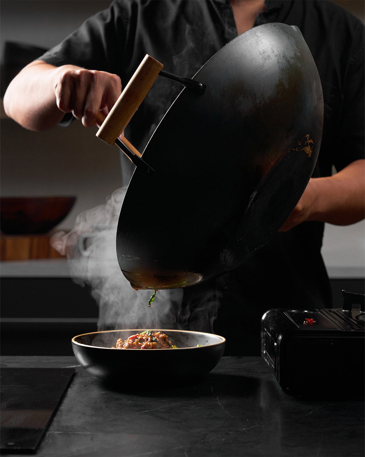 Chef plating hot stir-fry from a black steel wok into a bowl in a modern kitchen.