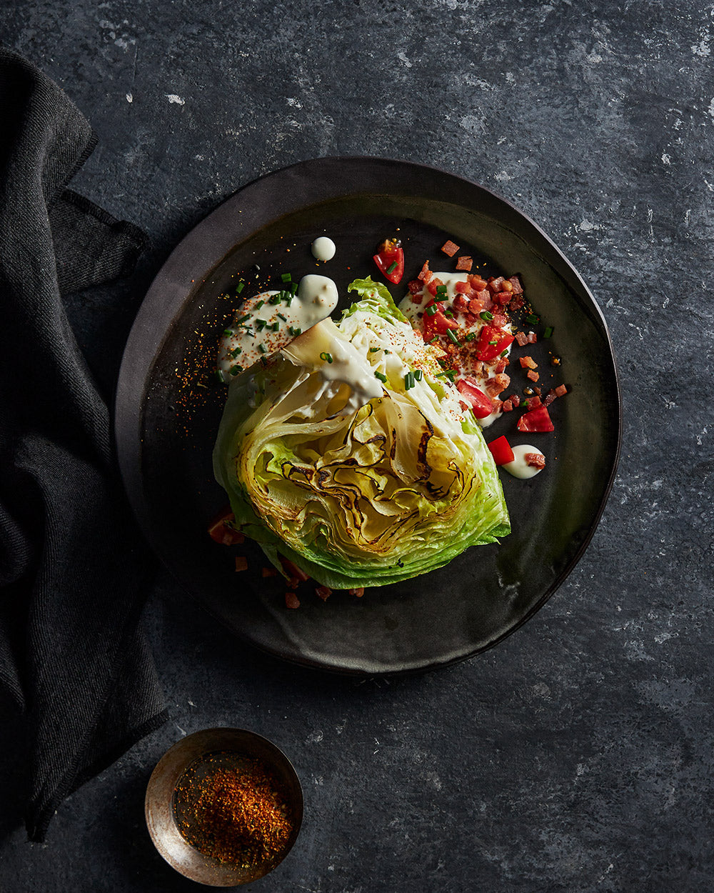 Charred iceberg lettuce wedge salad with diced tomatoes, crispy bacon, creamy dressing on black plate