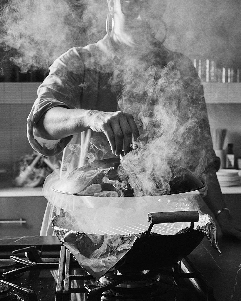 Person lifting smoky wok lid on gas stove in modern kitchen, black and white photo