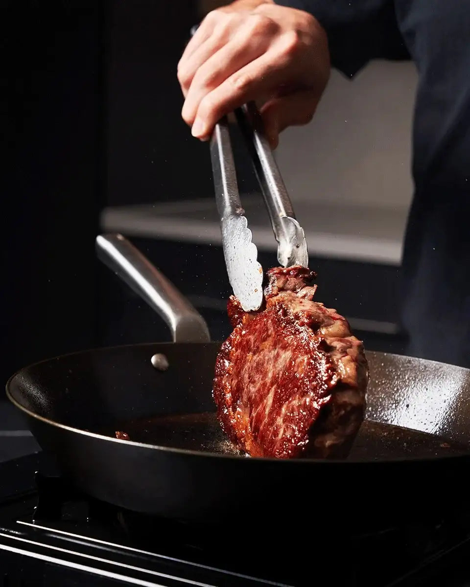 Hand using tongs to flip a seared steak in a black frying pan on stovetop, cooking process
